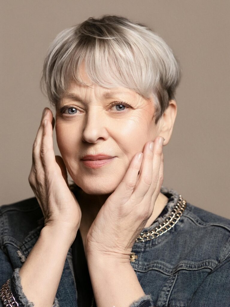 A woman with a smooth ash blonde pixie cut and full straight bangs that frame her blue eyes.