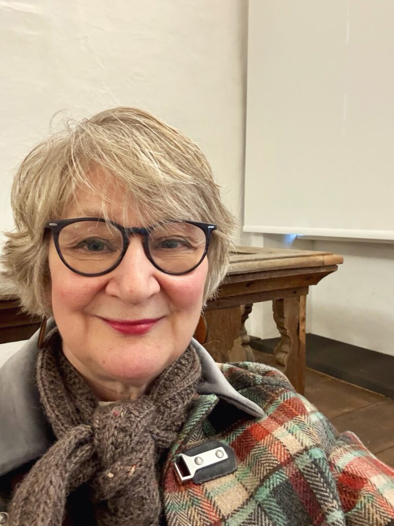 A smiling woman over 60 wearing glasses with an airy ash-blonde pixie cut and side-swept wispy bangs.