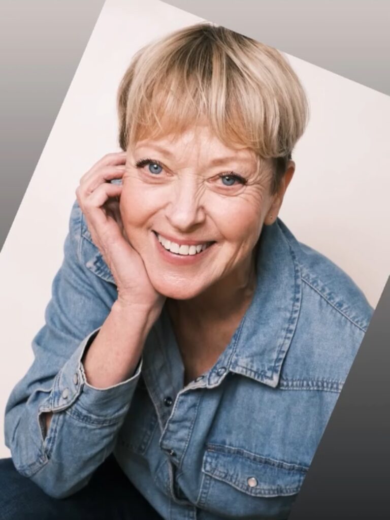 A woman over 60 with a short honey blonde pixie cut featuring choppy, piecey bangs and a denim shirt.