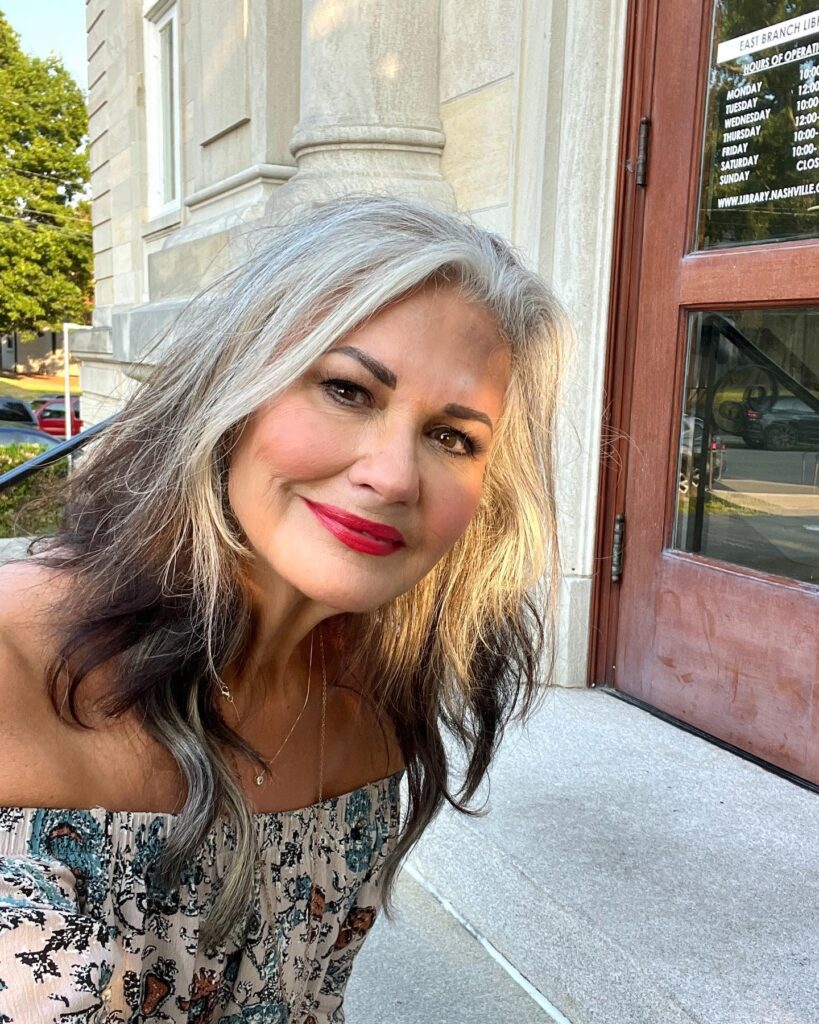 A woman with long wavy hair featuring a mix of dark charcoal and bright silver tones, styled with a side part and no bangs.
