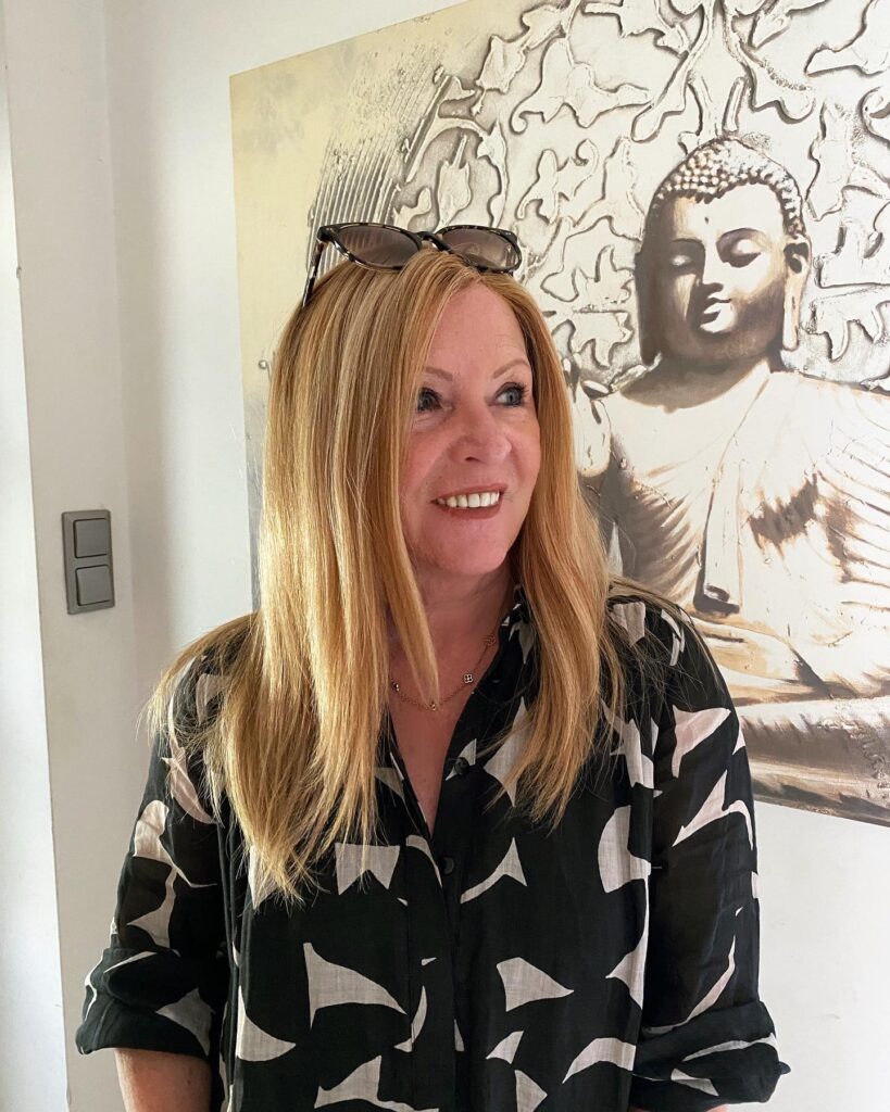 A woman over 60 with healthy honey blonde hair styled smooth past her shoulders without bangs.