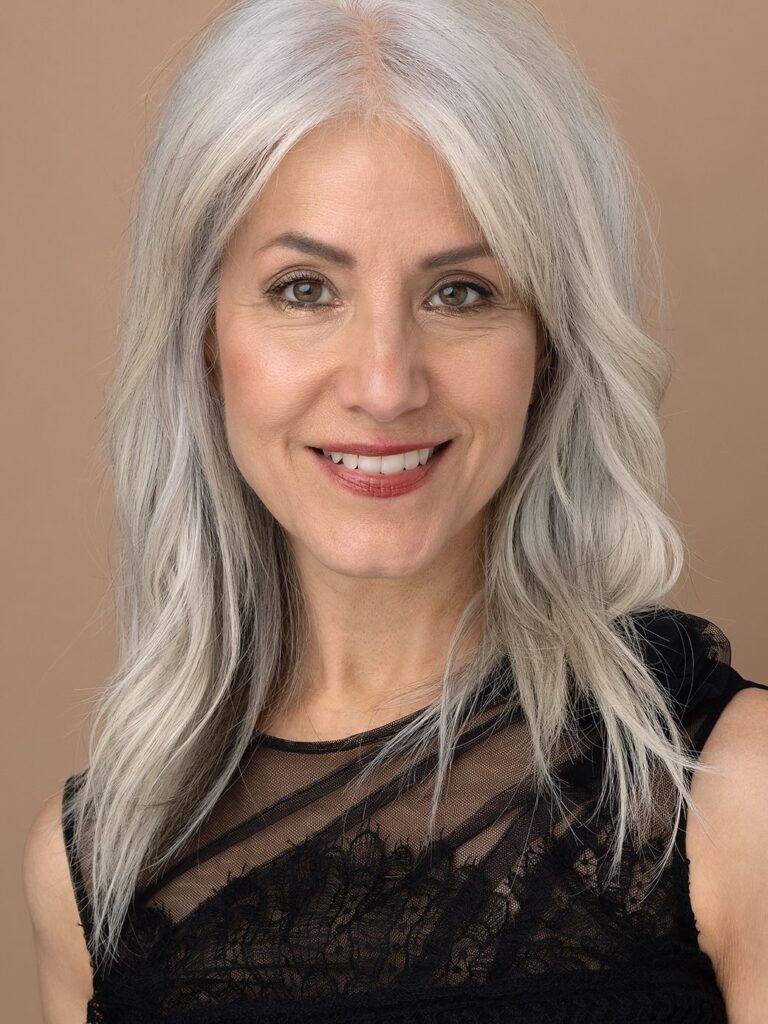 A middle-aged woman with shoulder-length, bright silver wavy hair that is parted in the middle with no bangs and styled with subtle layers.