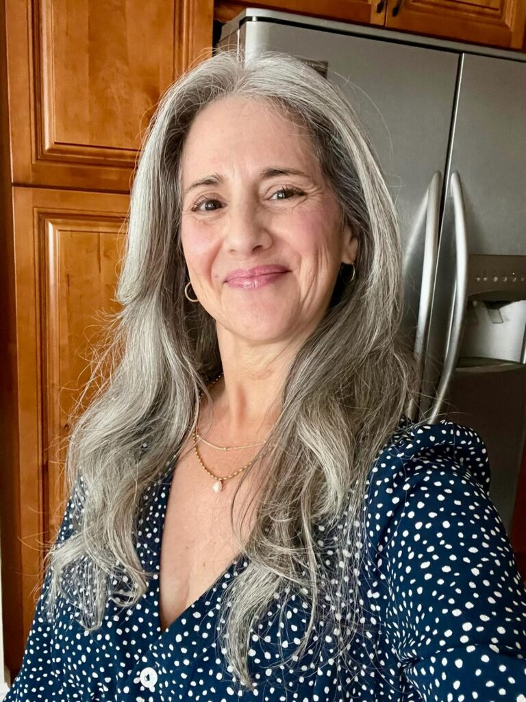 A woman with long, layered steel-grey hair styled in soft waves with no bangs.