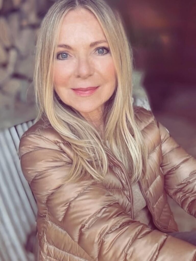A woman with long, straight golden-blonde hair featuring feathered ends and a middle part with no bangs.