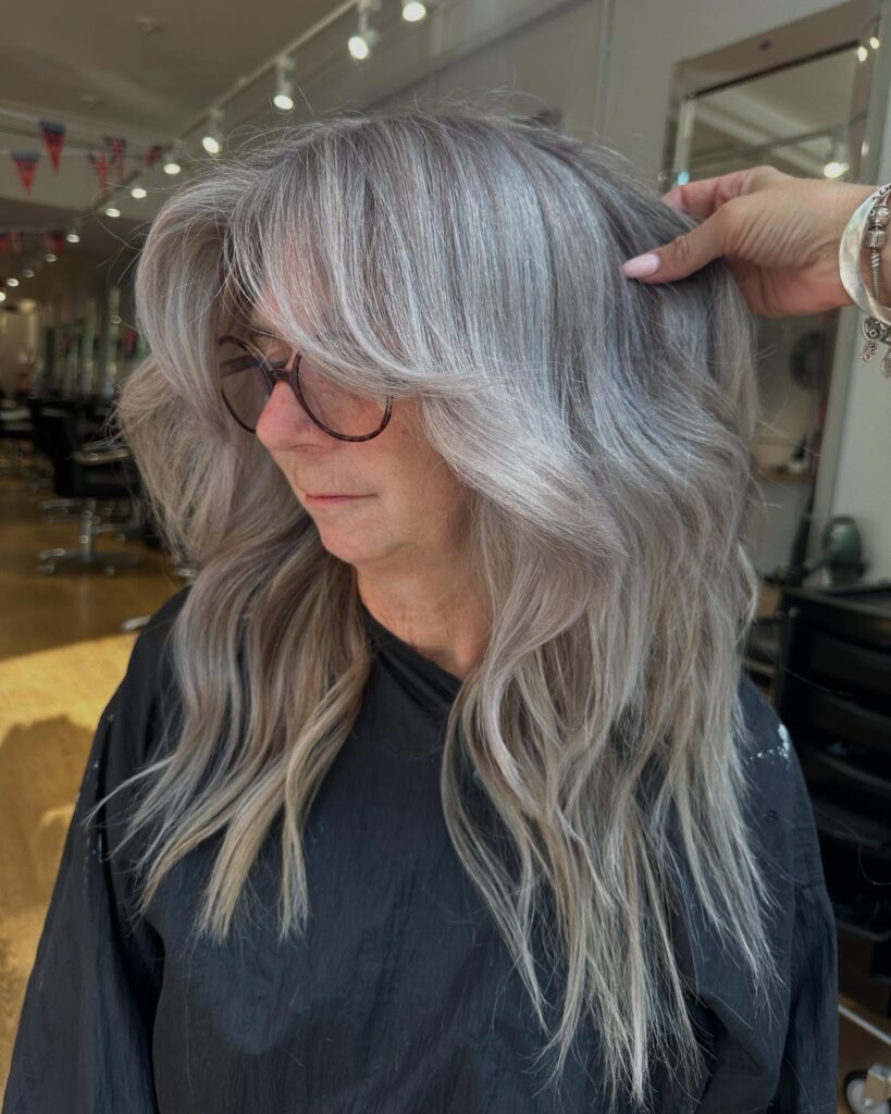 A woman with long, ashy silver hair styled in voluminous butterfly layers and a soft sweeping fringe.