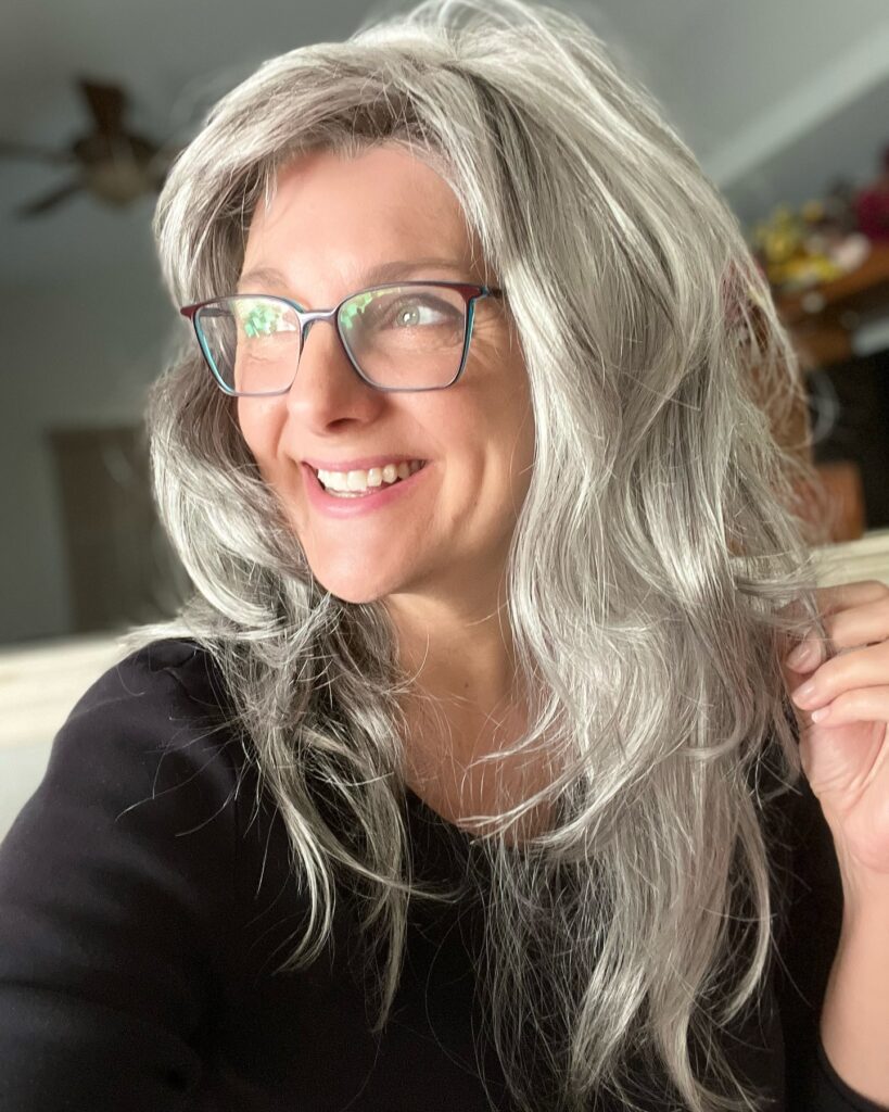 A smiling woman wearing glasses with long textured grey hair styled in piecey layers and wispy bangs.