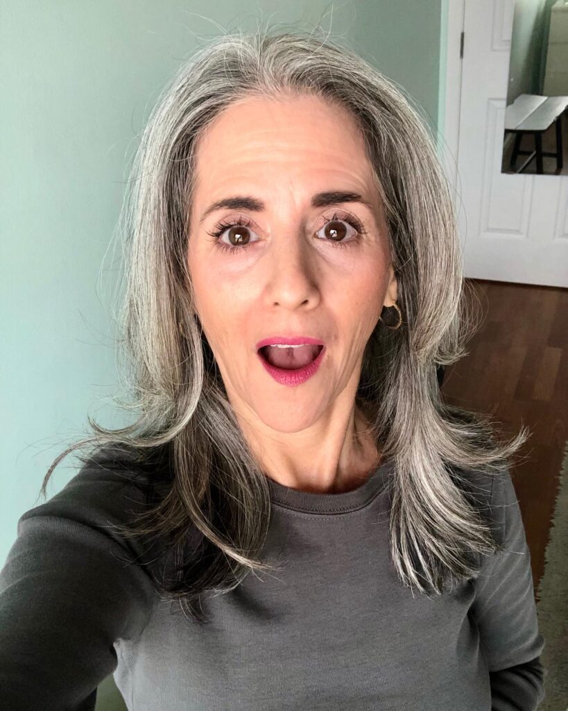 A woman with shoulder-length salt and pepper hair featuring soft layers and playful outward-flipped ends.