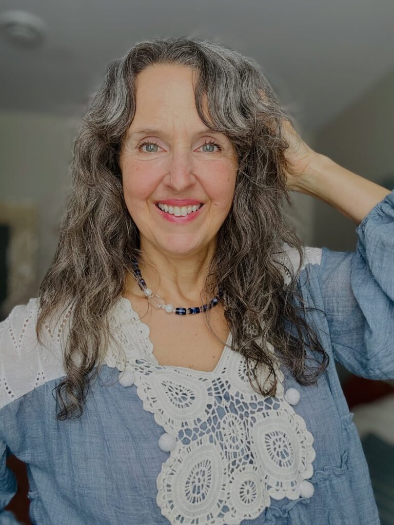 A smiling woman with long, curly salt-and-pepper hair and a soft, wispy fringe.