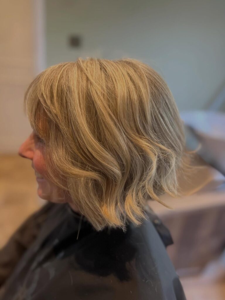A side profile of a woman over 60 with a neck-length choppy bob in caramel blonde with loose waves and textured ends.