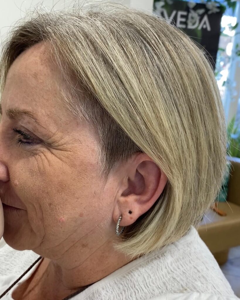 A side view of a woman over 60 with a straight blonde bob featuring a shaved undercut around the ear.
