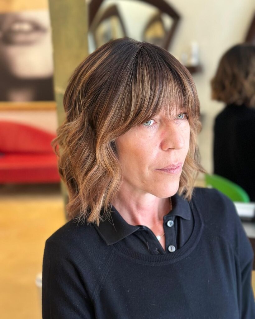 A woman over 60 with a shoulder-length wavy chestnut lob featuring caramel highlights and a textured fringe.