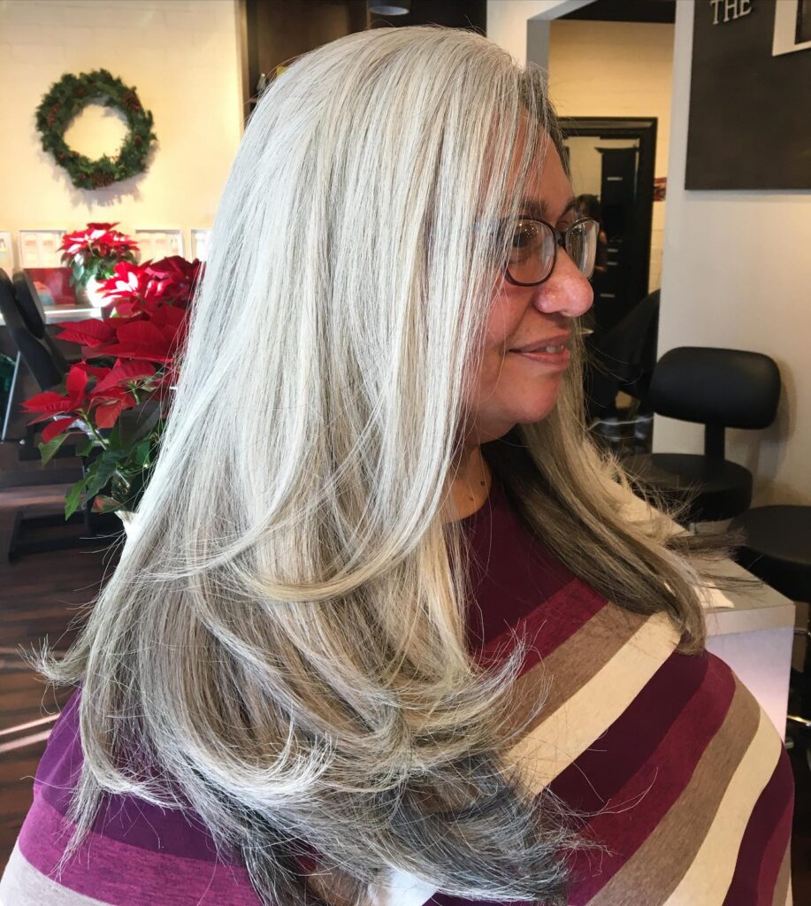 A side profile photo of a smiling woman with glasses and very long, natural silver hair cut with dramatic, cascading layers that are styled to curl slightly at the ends.