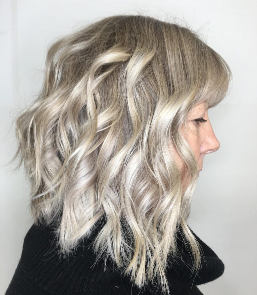 A side view of a woman with a shoulder-length wavy ash-blonde lob and soft wispy bangs.