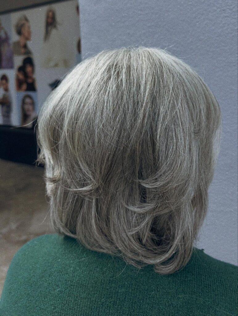 A back view of a woman with a neck-length salt-and-pepper bob featuring voluminous feathered layers flipped outward at the ends.