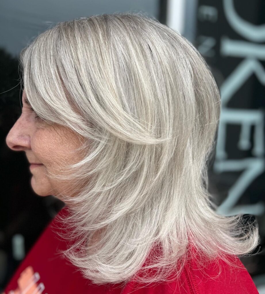 A side profile of a woman over 60 with medium-length silver hair featuring long, sweeping layers and feathered ends.