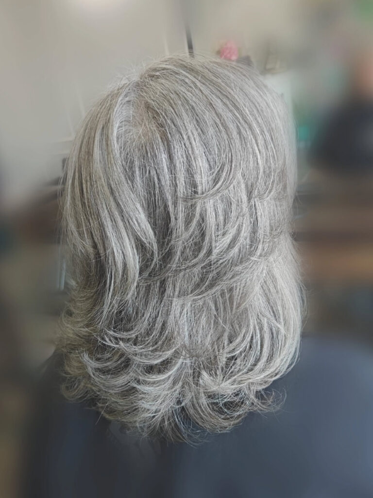 A back view of a woman over 60 with a medium-length feathered grey haircut featuring voluminous layers and textured ends.