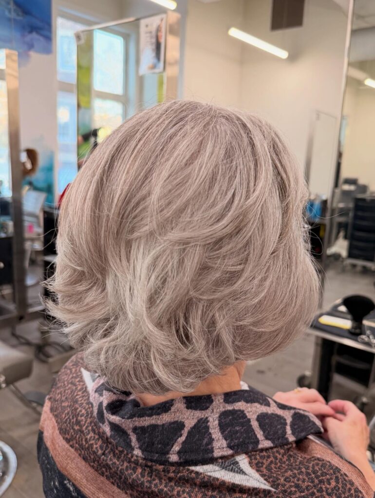 A back view of a woman with a neck-length layered hairstyle featuring a subtle dusty rose and silver color blend with textured feathered layers.
