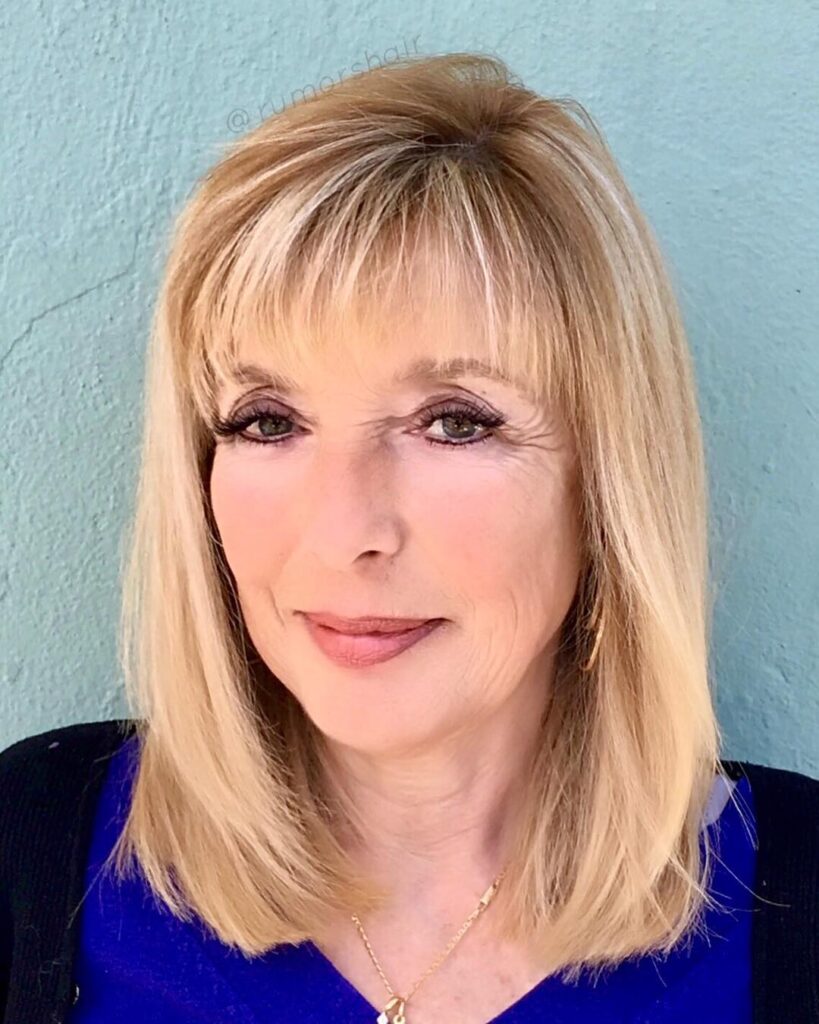 A front-facing photo of a woman with shoulder-length honey blonde hair featuring wispy bangs and soft layers.