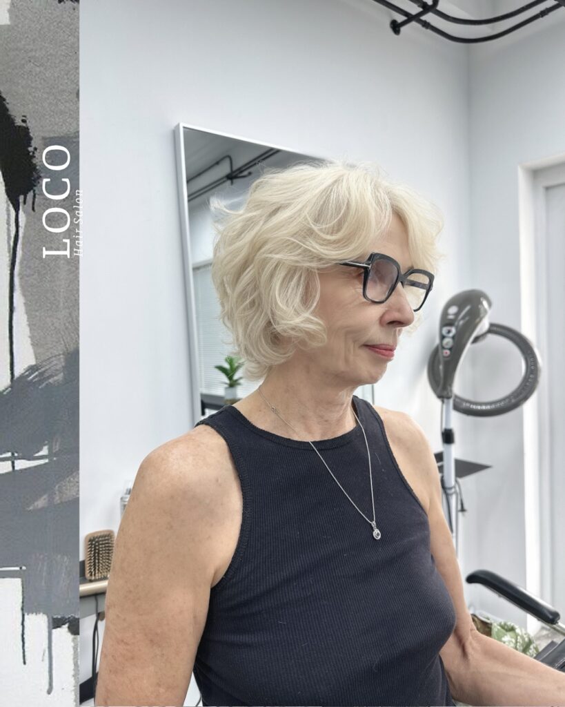A side profile of a woman with glasses and an ear-length platinum blonde layered shag with voluminous curls and wispy bangs.