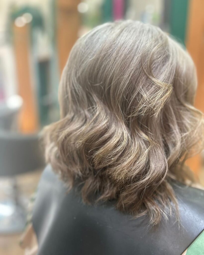 A back view of a woman over 60 with collarbone-length mushroom blonde hair styled in deep, voluminous waves.