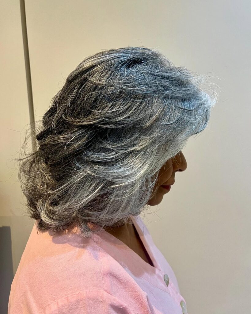 A side profile of a woman over 60 with a neck-length salt and pepper haircut featuring swept-back feathered layers.