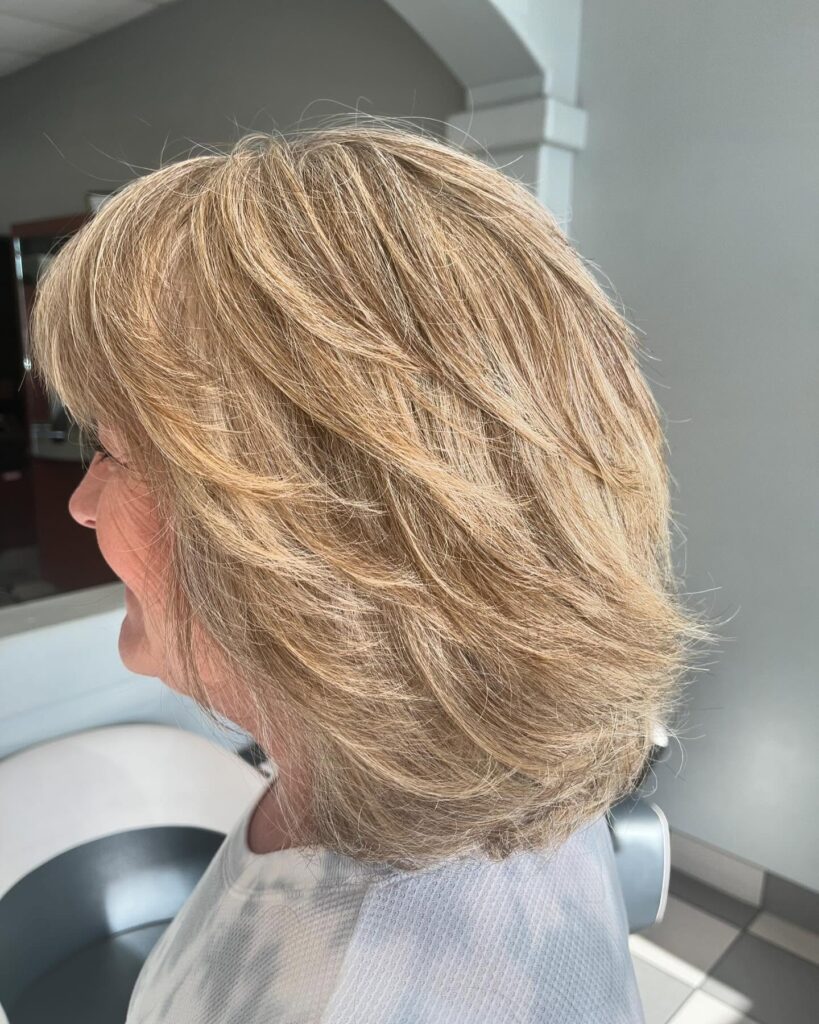 A side profile of a woman over 60 with a caramel blonde layered long bob that flows away from the face.