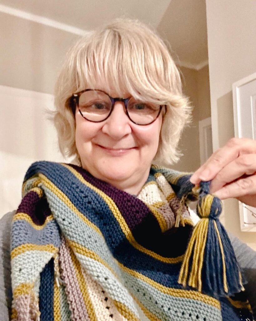 A woman with a chin-length buttery blonde shaggy bob and side-swept bangs, wearing glasses and a striped knit scarf.