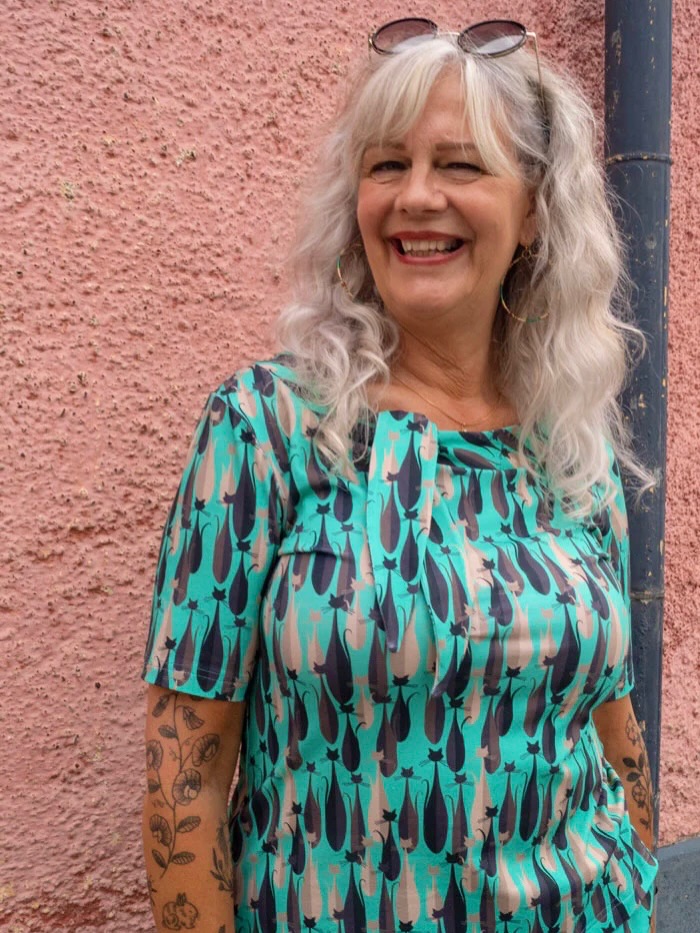 A smiling woman with long, wavy, icy silver hair and soft split bangs, wearing a teal cat-print top.