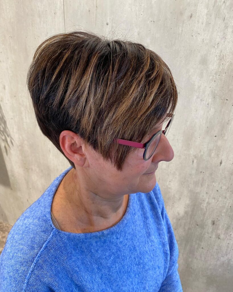 A woman over 60 with a short brunette asymmetrical pixie cut featuring honey-colored highlights and textured layers.