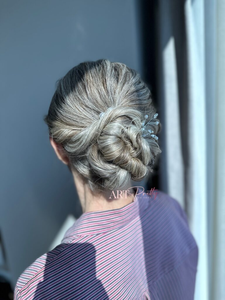 A woman with natural silver hair styled in a thick braided low bun adorned with a delicate pearl and crystal hairpiece.