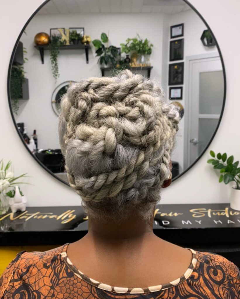 A woman with natural gray hair wears an elaborate updo consisting of thick rope twists woven into a decorative pattern.