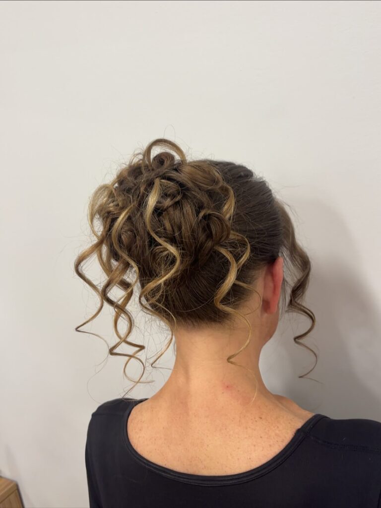 A woman is shown from the back wearing a high brunette updo with blonde highlights, and many defined ringlets cascading from the bun.