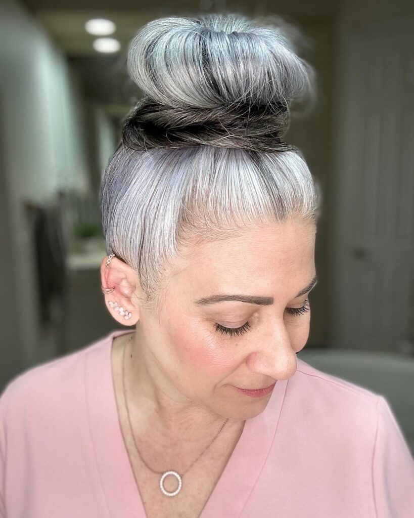 A woman with high-contrast silver and dark hair styled in a large, smooth topknot wrapped with a dark hair band.