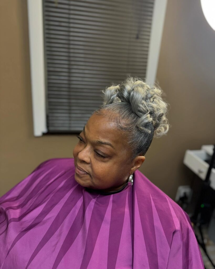 A woman wears a sleek, high updo featuring voluminous silver and blonde curls piled at the crown of her head.