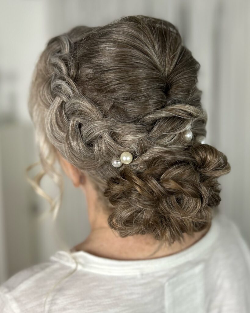A woman with highlighted brown hair styled in a side Dutch braid that leads into a low textured bun accented with pearl pins.