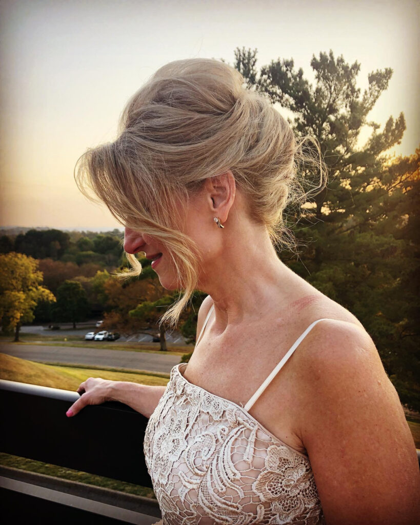 A woman with blonde hair is seen from the side, wearing a soft, voluminous French twist updo with loose, wavy strands framing her face.