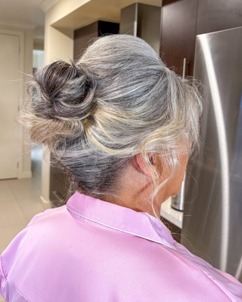 A woman with wavy silver and gray hair is shown from the back, wearing a casual messy bun with loose strands framing her neck.