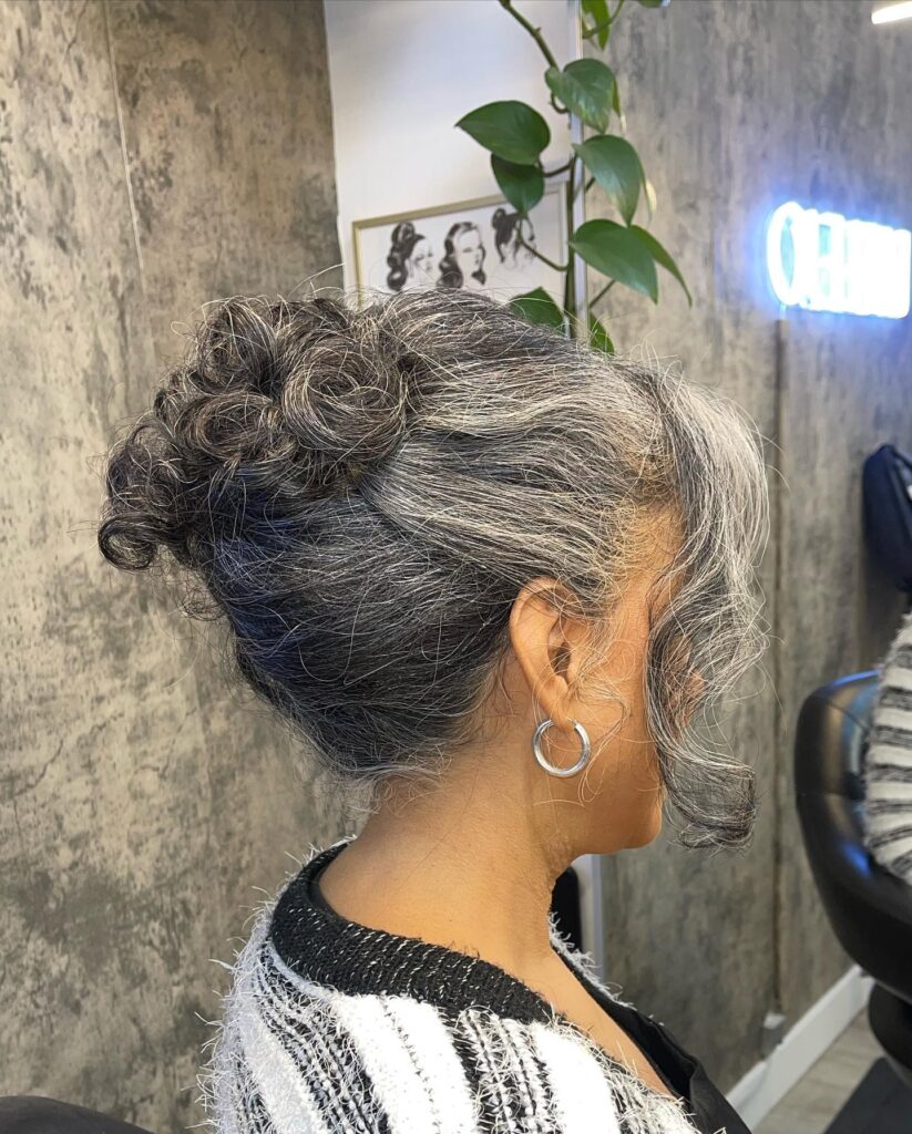 A woman with curly salt-and-pepper hair styled in a back up-do of small rosettes with long wavy bangs.