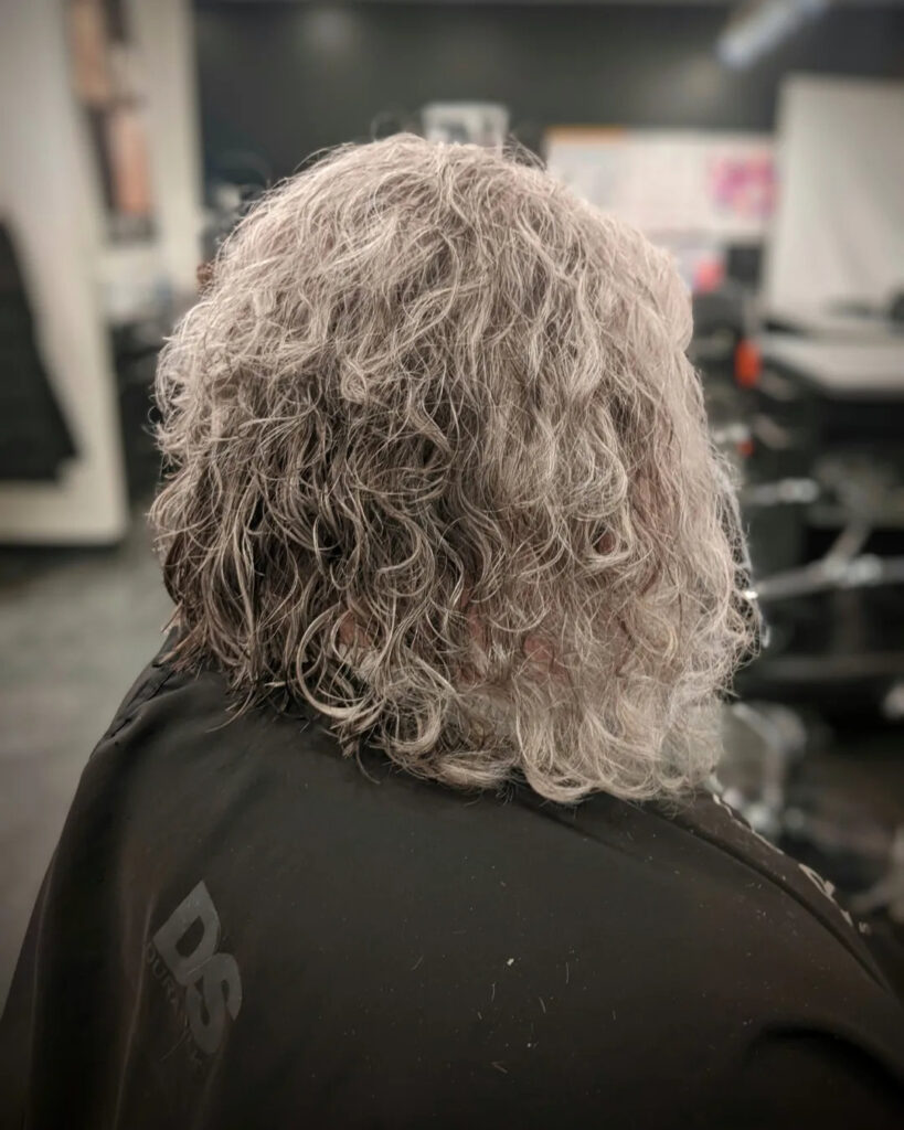 A back view of a woman with a curly salt-and-pepper inverted bob showing lots of natural volume and texture.