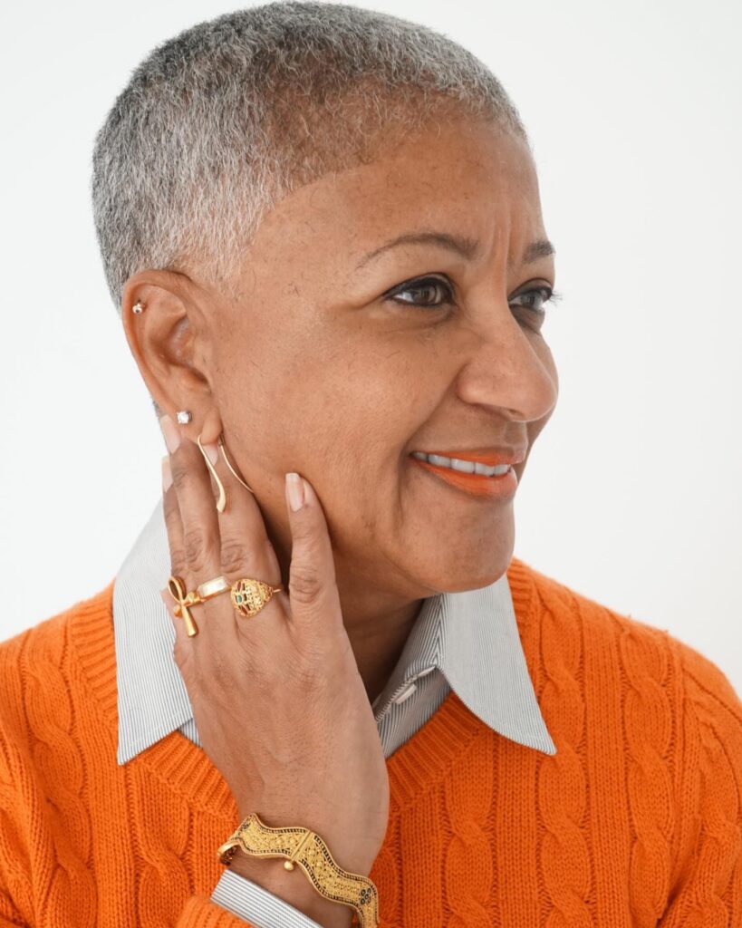 A woman with a very short silver buzz cut smiling while wearing an orange sweater and gold jewelry.