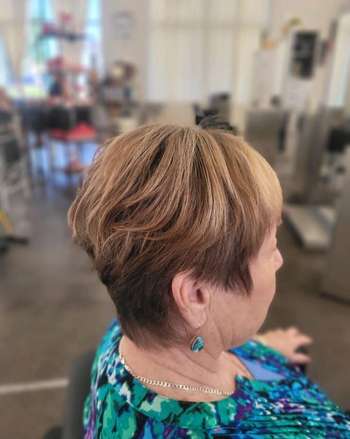 A woman viewed from the side, showing a stacked honey blonde pixie haircut with short layers at the back.