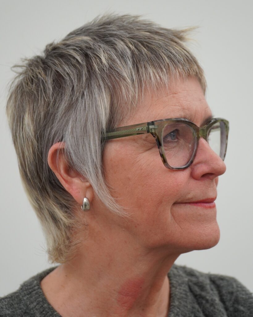 A woman with a wispy ash blonde pixie cut featuring very short bangs and feathered side layers.