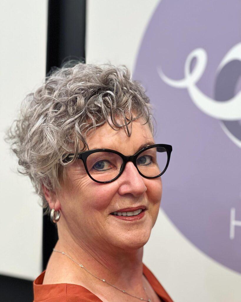 A woman over 60 with a voluminous curly salt-and-pepper pixie haircut and black-frame glasses.