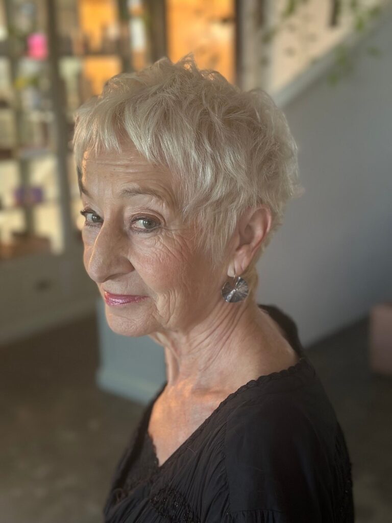 A woman with a textured champagne blonde pixie cut featuring short wispy bangs and ruffled layers on top.