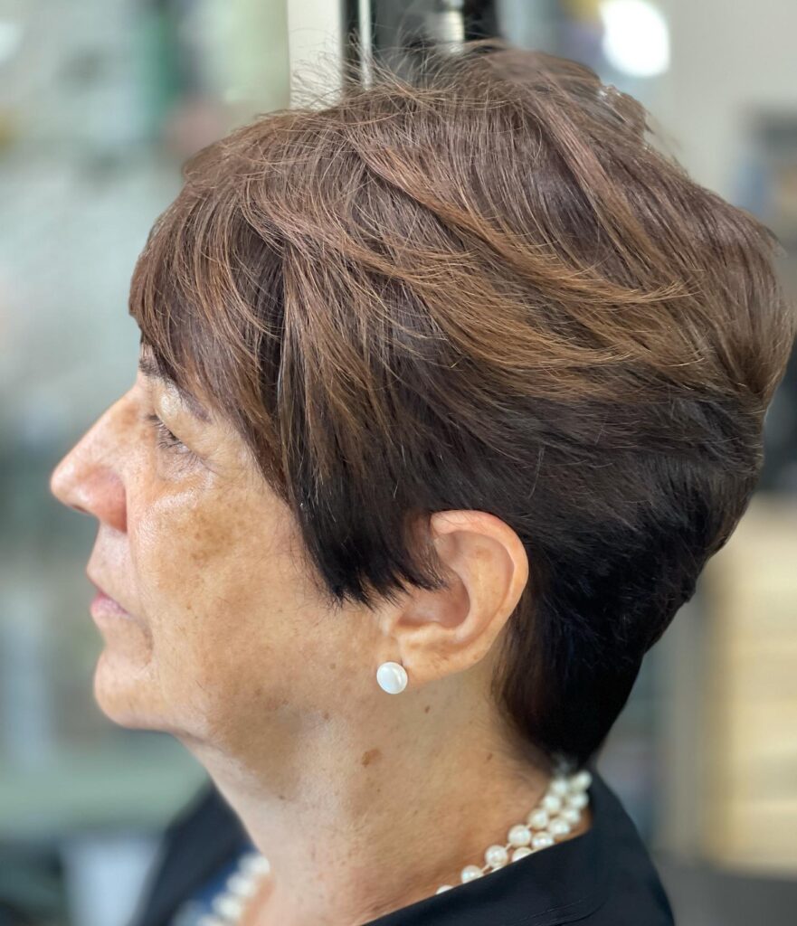 A woman in profile with a chestnut brown pixie haircut featuring voluminous layers swept back from the forehead.