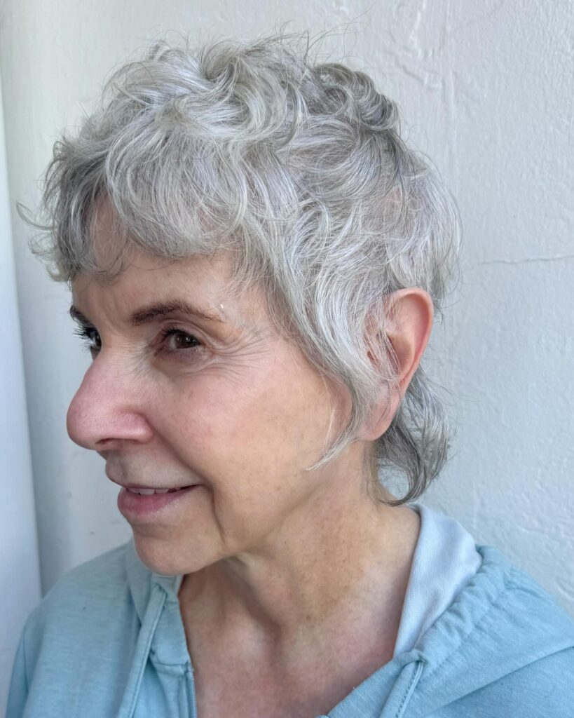 A woman over 60 with a wispy silver pixie haircut featuring wavy layers and soft tapered edges.