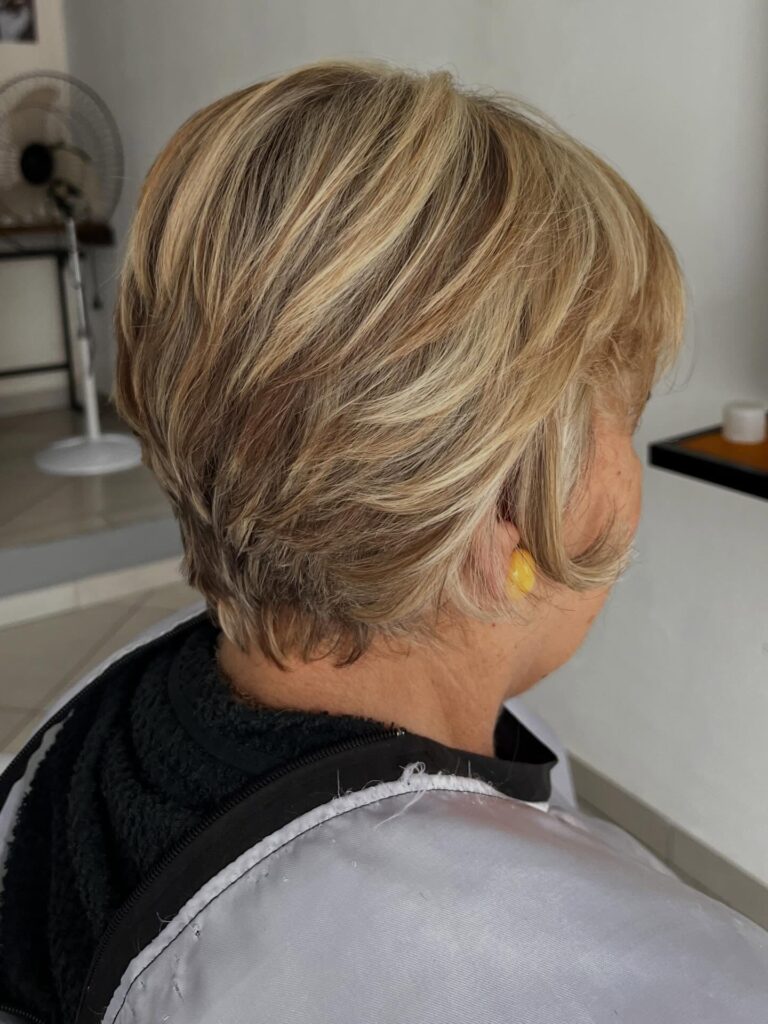 A woman over 60 with a layered honey-blonde pixie haircut featuring multi-dimensional highlights and feathered texture.