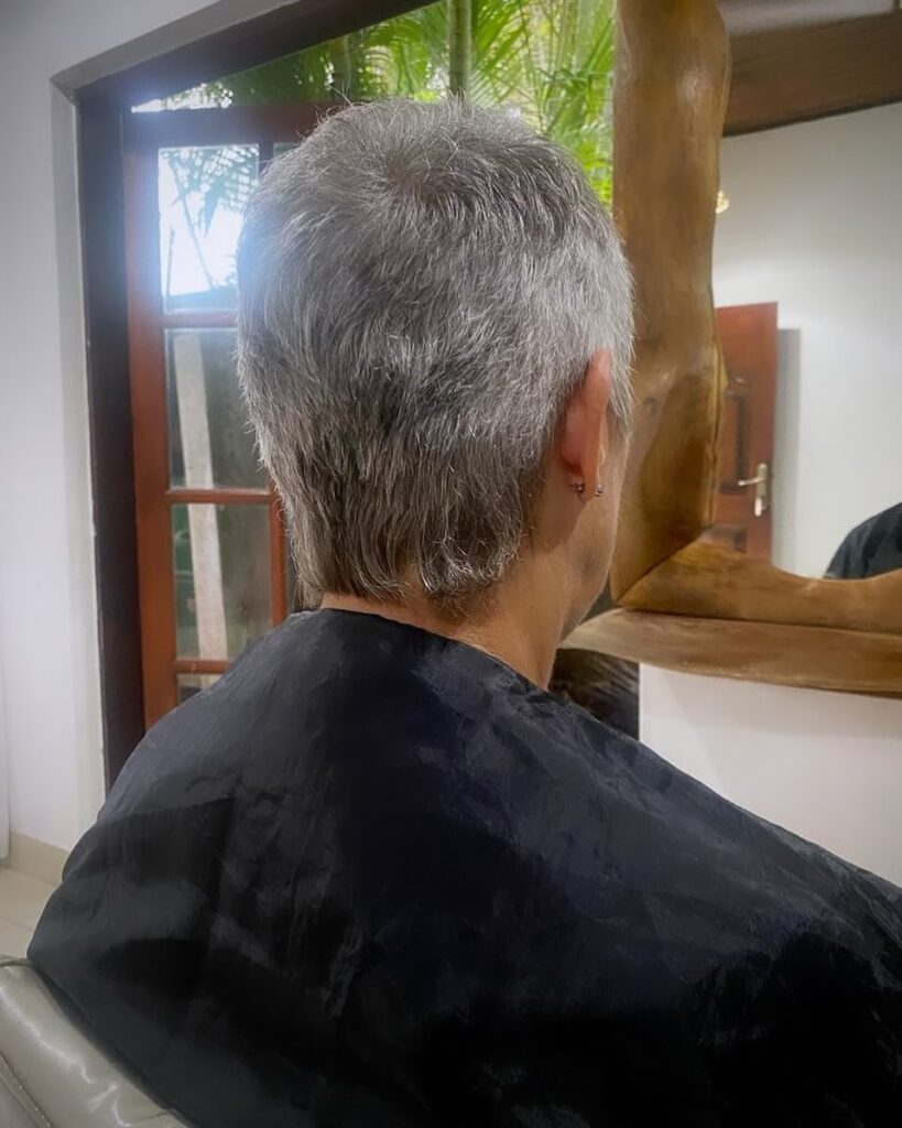 A rear view of a woman with a natural grey pixie haircut featuring soft feathered layers and a slightly longer, wispy nape.