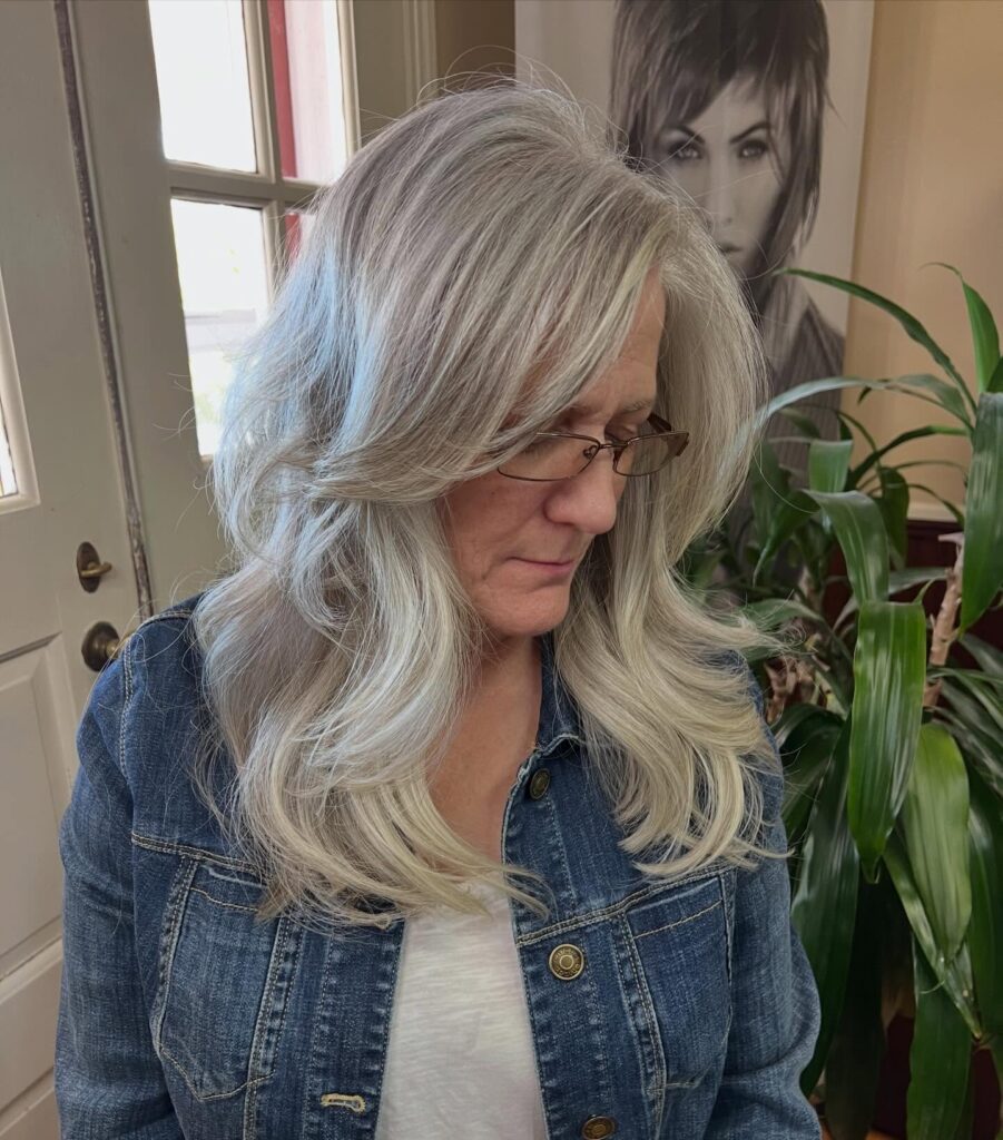 A woman with long, layered silver hair featuring a deep side part and soft curls at the ends.