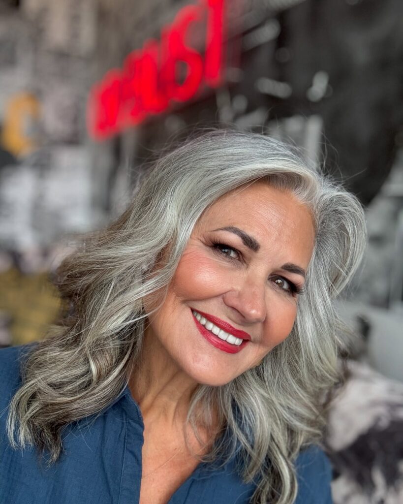 A smiling woman over 60 with a round face and voluminous medium-length silver hair styled in soft waves with a deep side part.